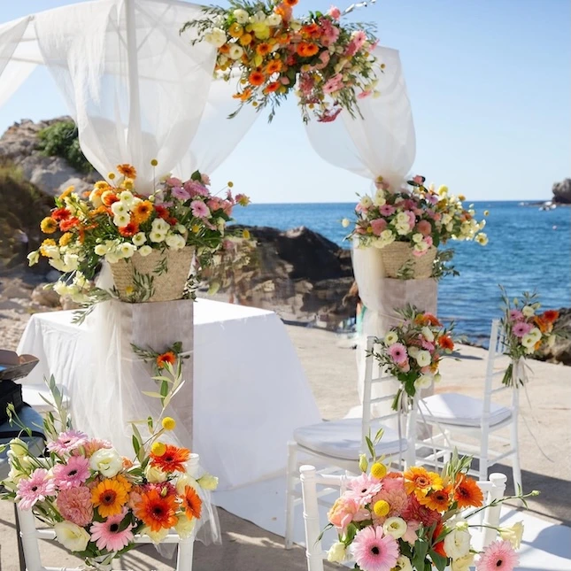 Un mariage d’exception organisé dans l'esprit de Mondello Hospitality dans une baie secrète et entièrement privatisée du Sud de la France, entre mer turquoise et falaises méditerranéennes. La cérémonie laïque se déroule en plein air, sous un autel élégant décoré de voilages blancs et d’une arche florale foisonnante inspirée des couleurs du Sud, dans des tons solaires : jaune citron, rose, orange et blanc. Une allée centrale recouverte d’un tapis blanc, bordée de pétales, mène à l’autel. Les invités sont confortablement installés sur des chaises Napoléon blanches, parfaitement alignées pour une cérémonie raffinée et structurée. L’ensemble crée un cadre intime, poétique et résolument élégant, à l’image des criques sauvages de la Côte d’Azur et des origines franco-sicilienne de Mondello