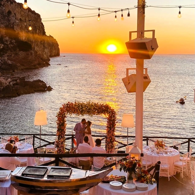 Réception les pieds dans l’eau au coucher du soleil – ambiance Dolce Vita dans une baie secrète organisée par Mondello Hospitality. Après la cérémonie, les invités découvrent une terrasse surplombant la mer, idéale pour un cocktail et un dîner festif dans un esprit chaleureux et méditerranéen. Guirlandes lumineuses, lampes d’ambiance, compositions florales généreuses, nappes blanches et tables dressées avec raffinement : tout est réuni pour une réception privée au bord de l’eau, sublimée par un coucher de soleil magique. L’atmosphère rappelle les plus beaux soirs d’été sur la Riviera, entre authenticité et élégance naturelle. Un WOW dans l'esprit de Mondello Hospitality